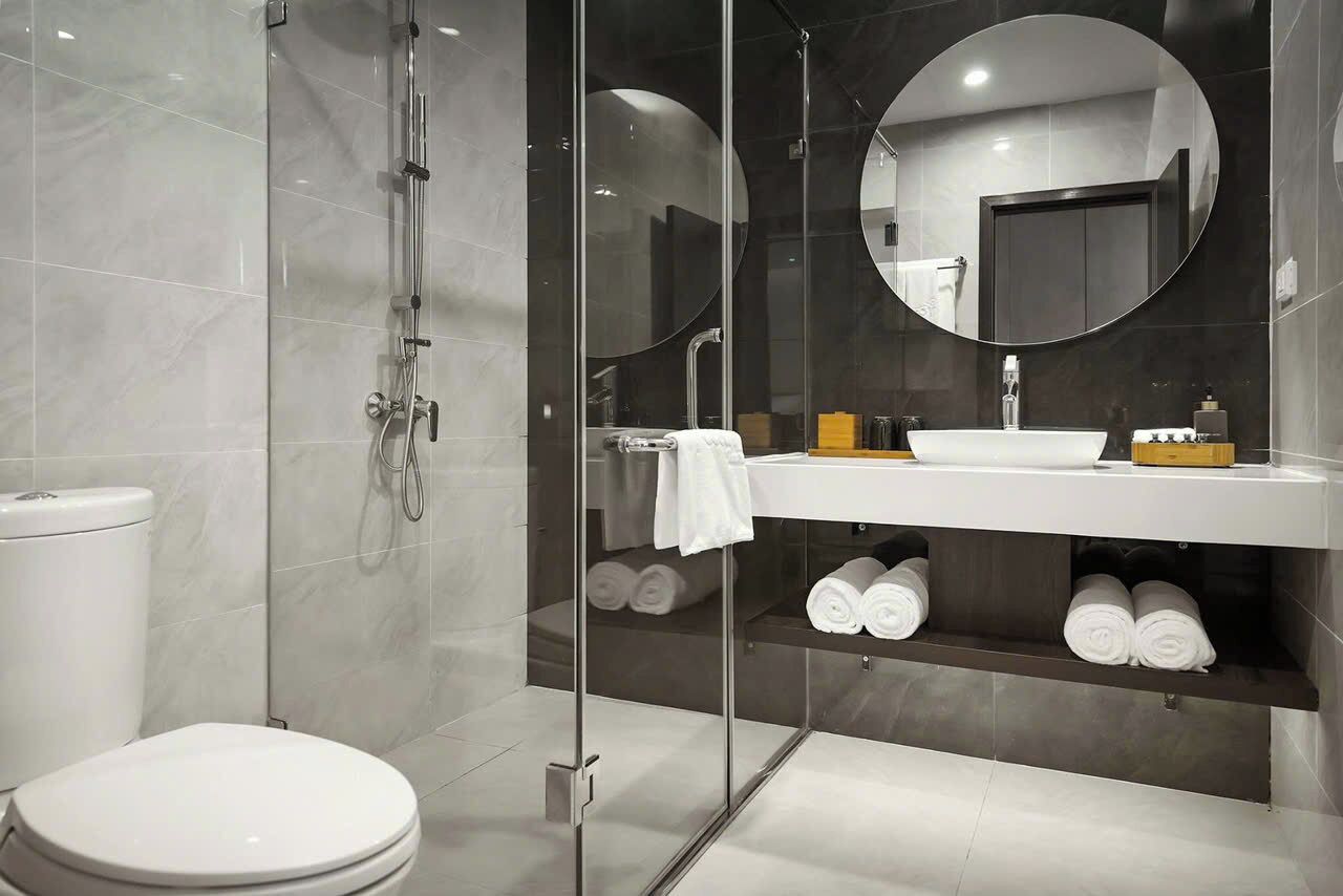 Un baño moderno con ducha envuelta en vidrio, inodoro blanco, espejo redondo y lavamanos blanco con perchero.