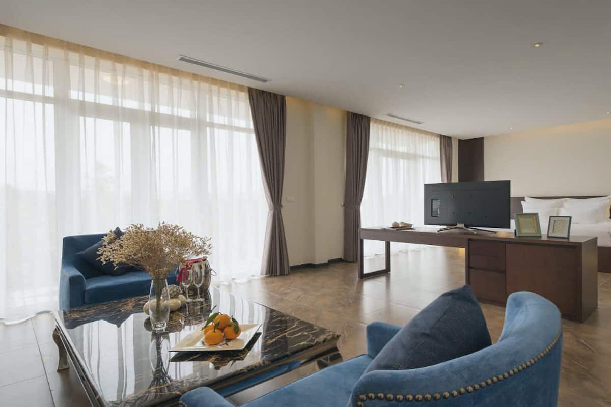 Een moderne hotelkamer met een groot bed, een bureau met een flatscreen televisie, een bank en een glazen salontafel met decoratieve objecten.