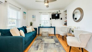 Un salon avec un canapé bleu, un fauteuil beige, une table basse, un téléviseur fixé au mur et un ventilateur de plafond.