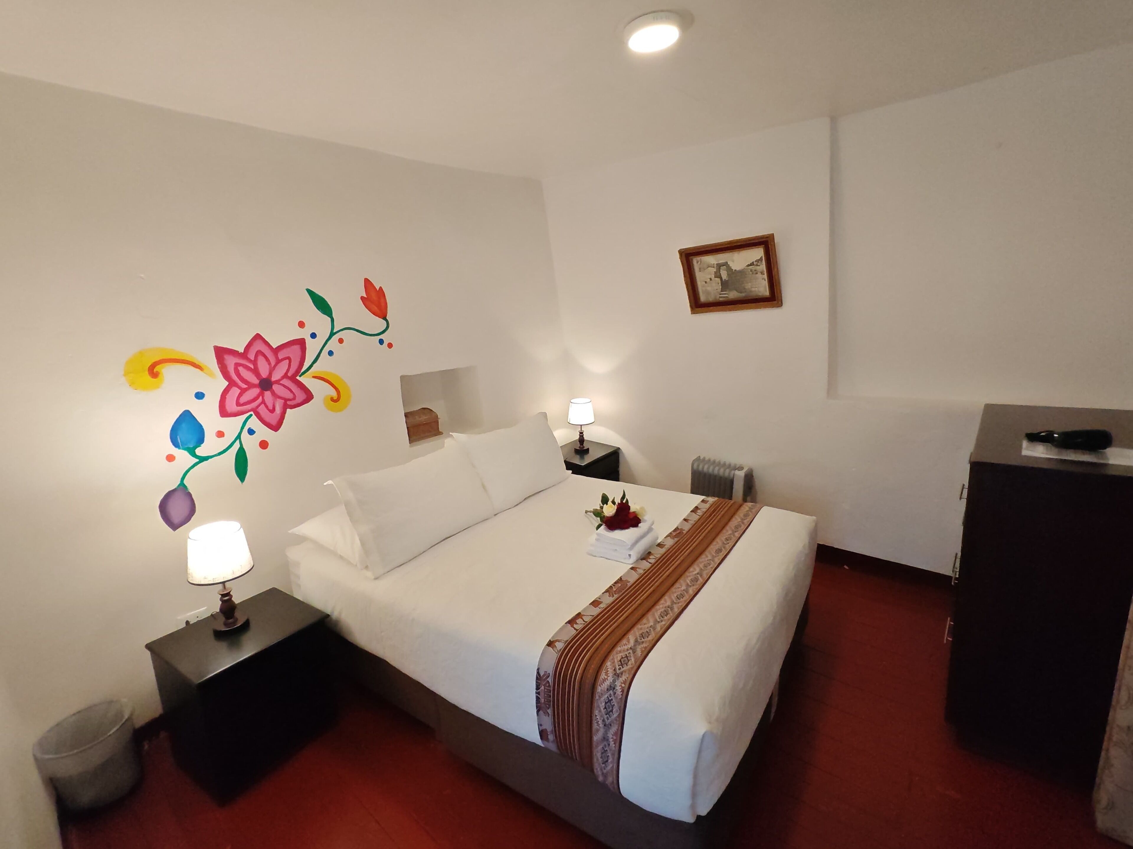 A hotel room with a bed, bedside tables, a dresser, a radiator, and a wall with floral artwork.