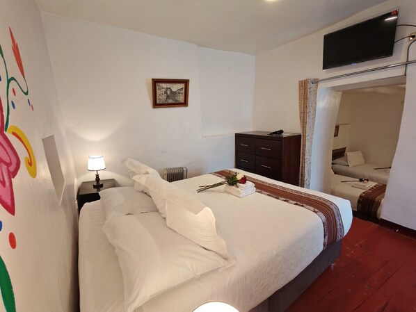 Habitación de hotel con una cama bien hecha, una lámpara de noche, un cuadro enmarcado en la pared, un aparador de madera oscura y un televisor montado.