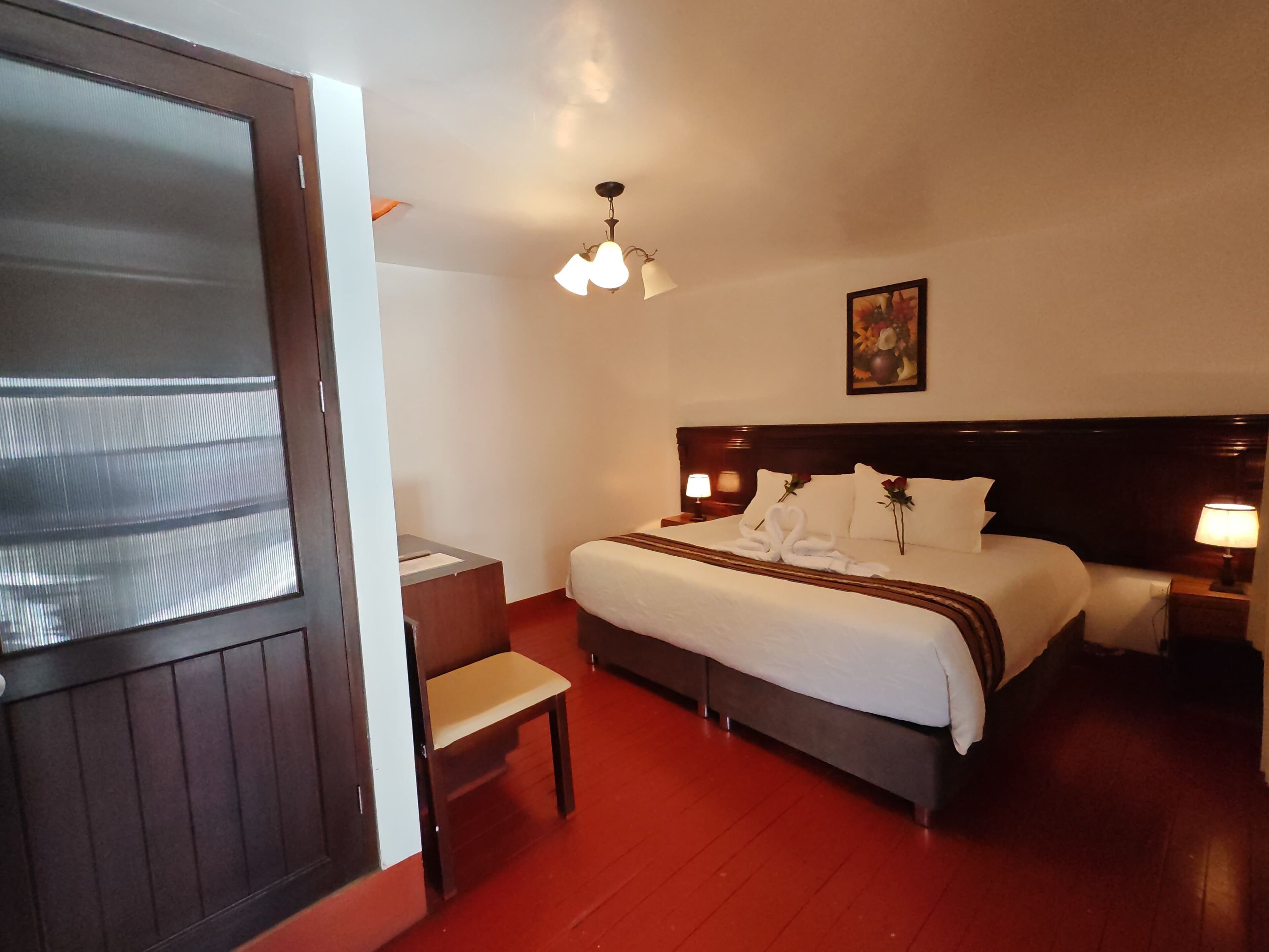 A hotel room with a bed, bedside tables, a desk, a chair, a painting on the wall, and a ceiling light.