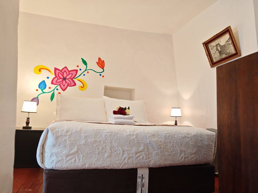 Una cama bien hecha con un adorno floral en la pared, dos lámparas de noche y un cuadro enmarcado.