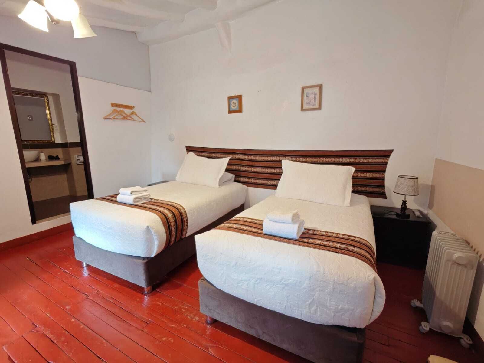 Habitación de hotel con dos camas, cabecera de madera, una mesita de noche con lámpara y un radiador.