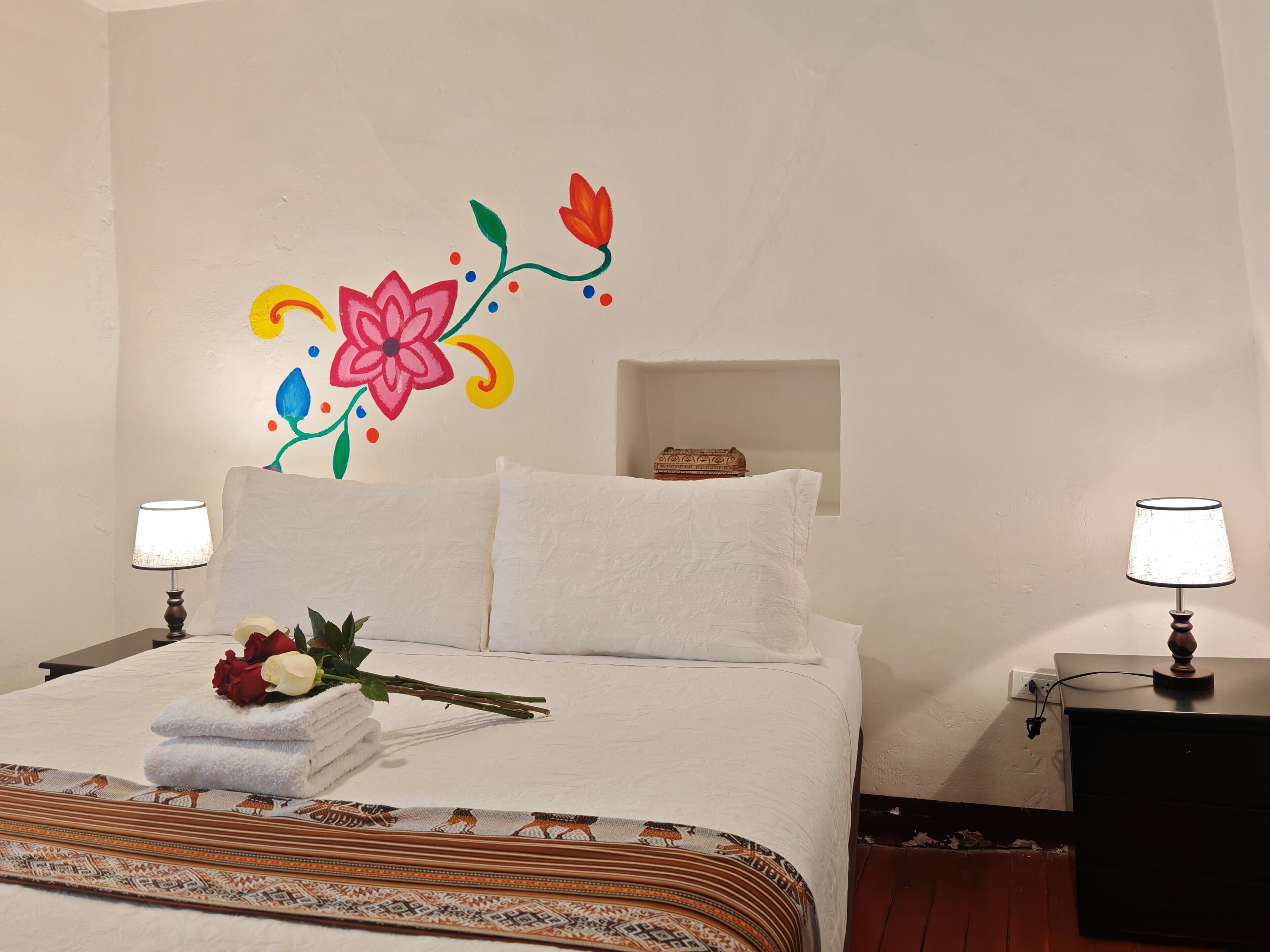 Una cama bien hecha con sábanas blancas, un adorno floral en la pared y una mesita de noche con lámpara.