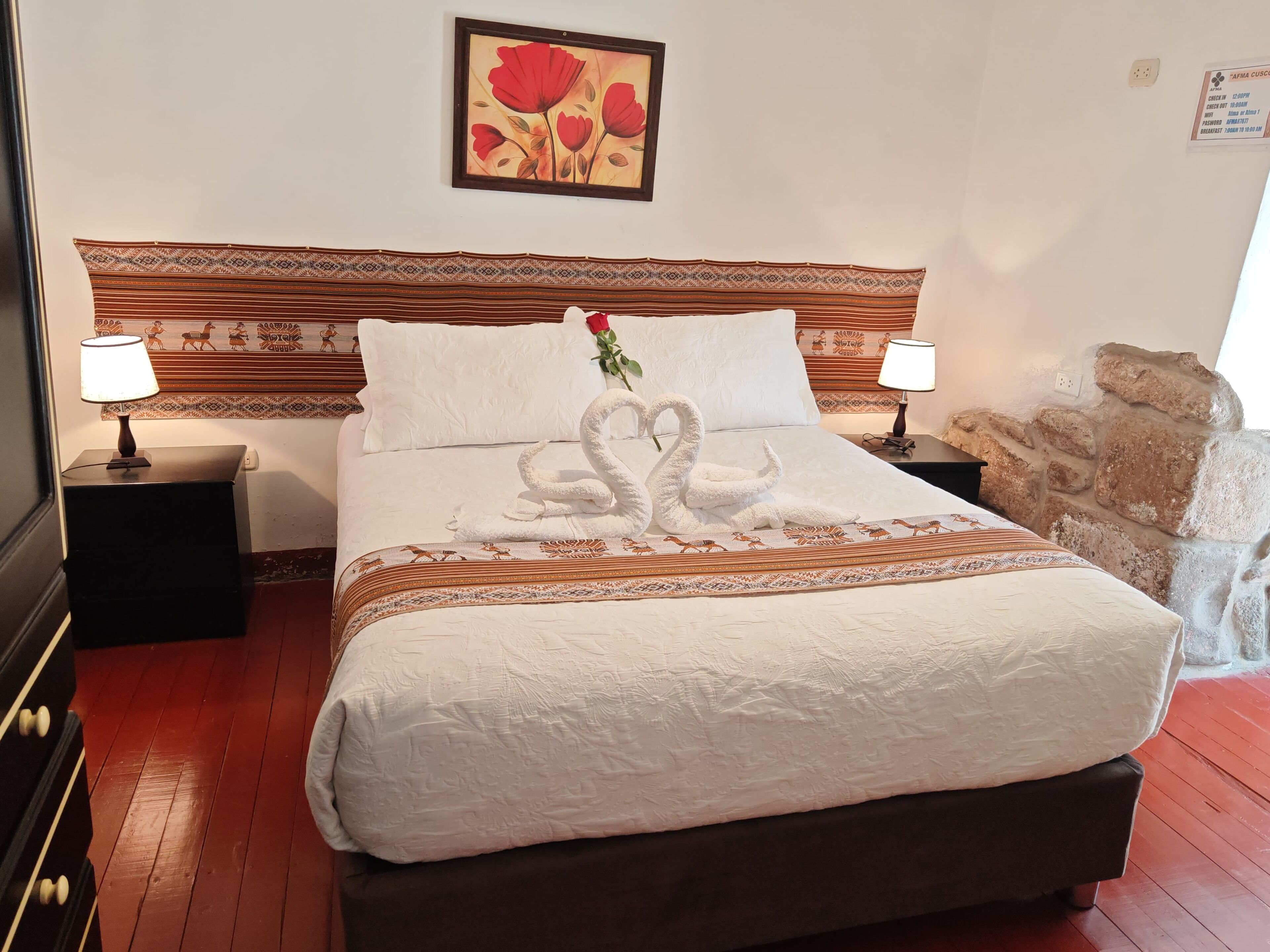 A neatly made bed with a white duvet, two bedside lamps, a decorative headboard, a framed artwork above the bed, and a stone wall feature to the right.