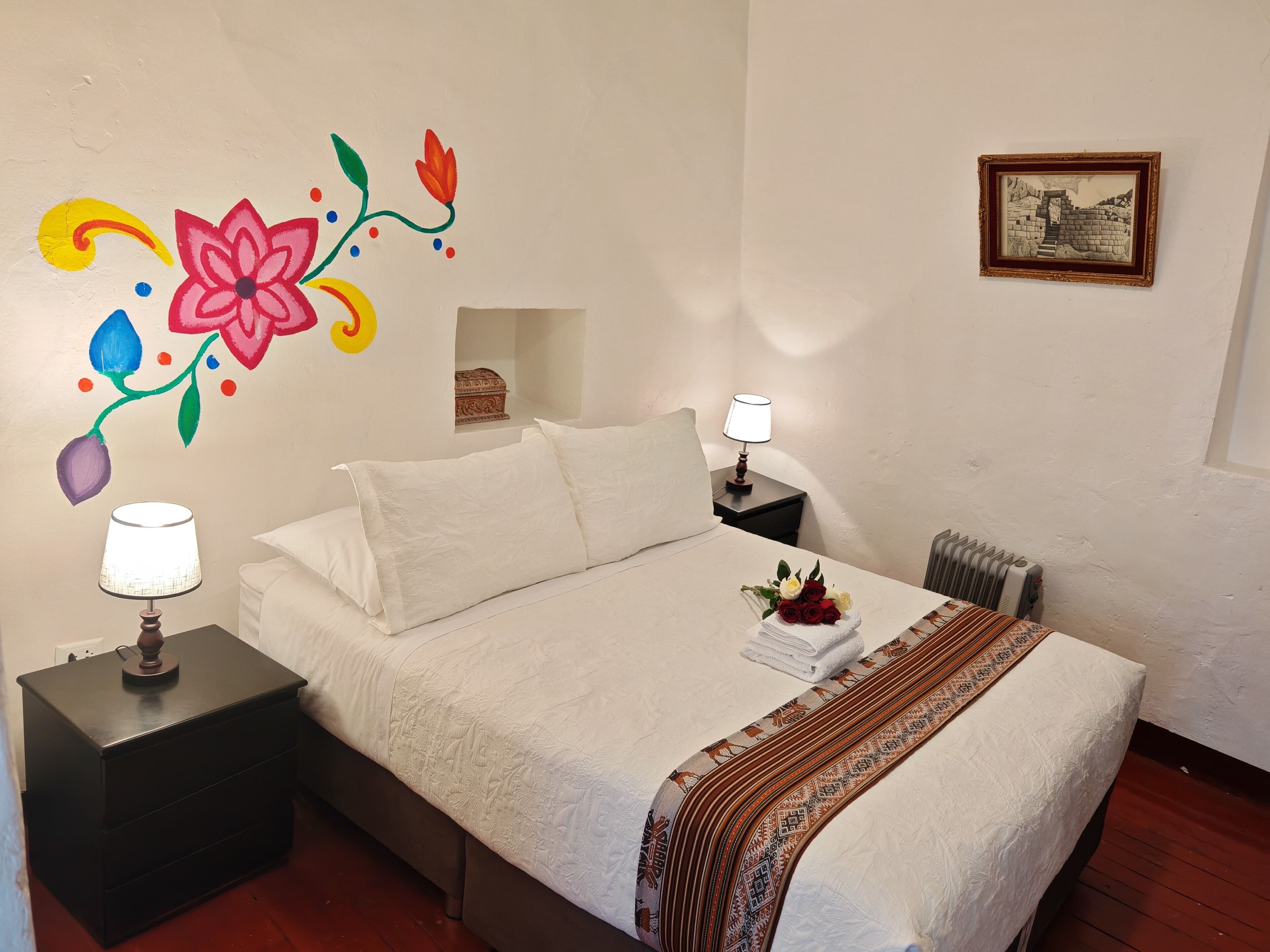 A bedroom with a bed, two bedside tables, a lamp, a radiator, a wall decoration, and a framed picture.