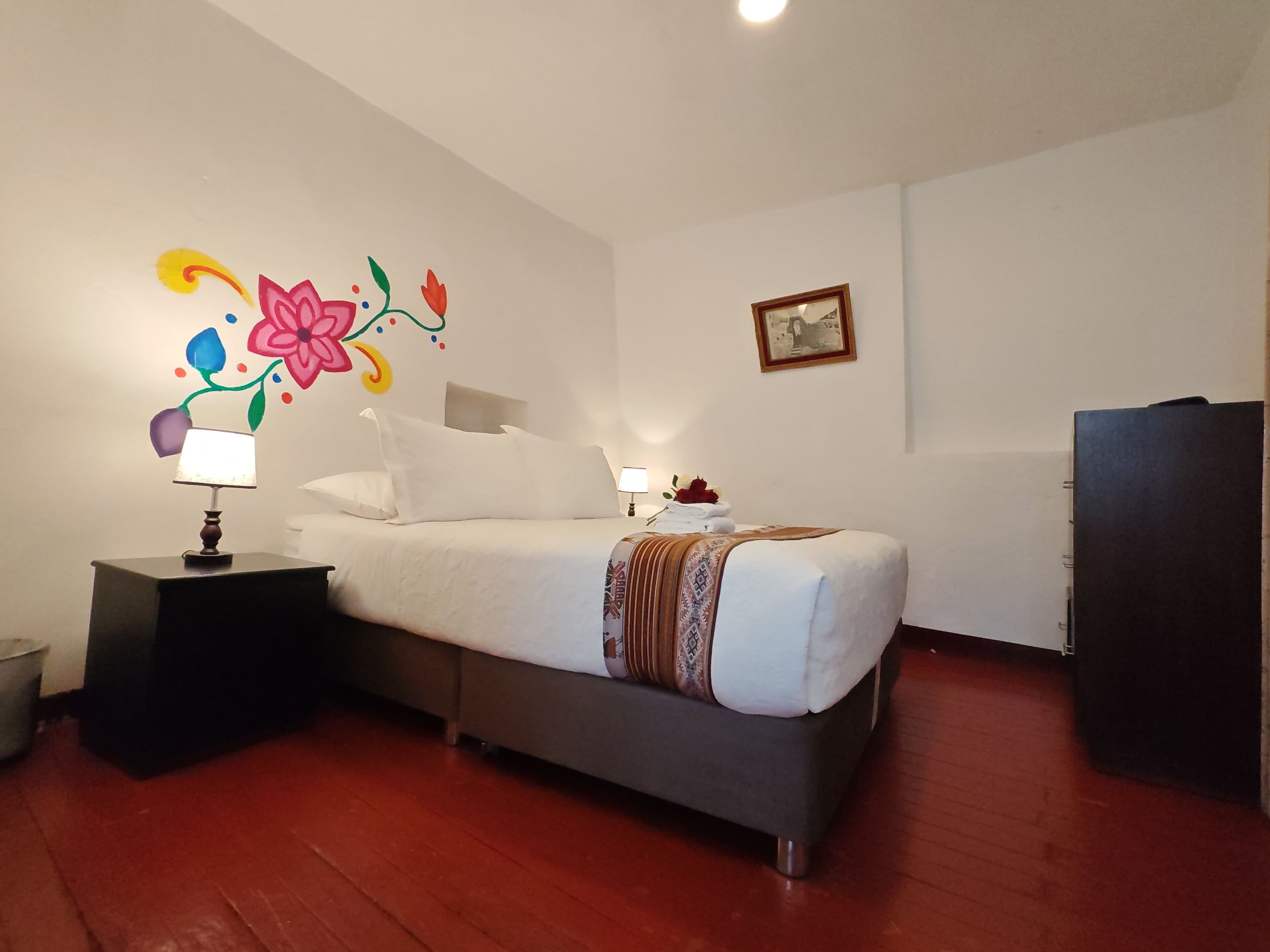 A neatly made bed with a white comforter and pillows, a bedside lamp, a decorative wall mural, a framed picture, and a dark-colored wardrobe.