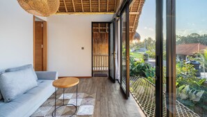 A modern living room with a sofa, a round wooden table, and a large glass door leading to a balcony with a view of greenery and a house. - Green Harmony Villa by Betterplace (Ubud)