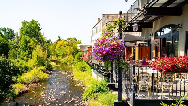 Un café junto al río con terraza, un puente de piedra y abundante vegetación.