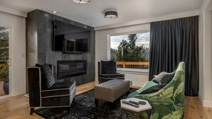 Un salón moderno con chimenea, muebles negros y verdes, y un ventanal con vistas a árboles.