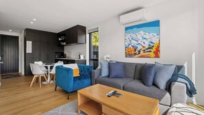 Una sala moderna con un sofá azul, un sofá gris y una mesa de centro de madera. Se ve una cocina al fondo con armarios oscuros y una ventana.