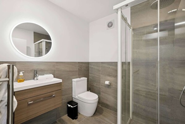 Un baño moderno con cabina de ducha de vidrio, espejo redondo, inodoro blanco y tocador de madera con lavamanos y dispensador de jabón.
