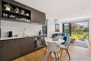 Una cocina moderna con armarios oscuros, azulejos tipo metro blancos y un horno empotrado. Hay un comedor con una mesa redonda y sillas, y un ventanal con vistas a un jardín y una casa.