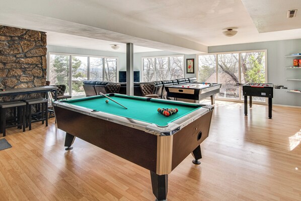 Une table de billard dans une pièce avec un mur en pierre, un coin bar et un espace salon offrant une vue sur les arbres.