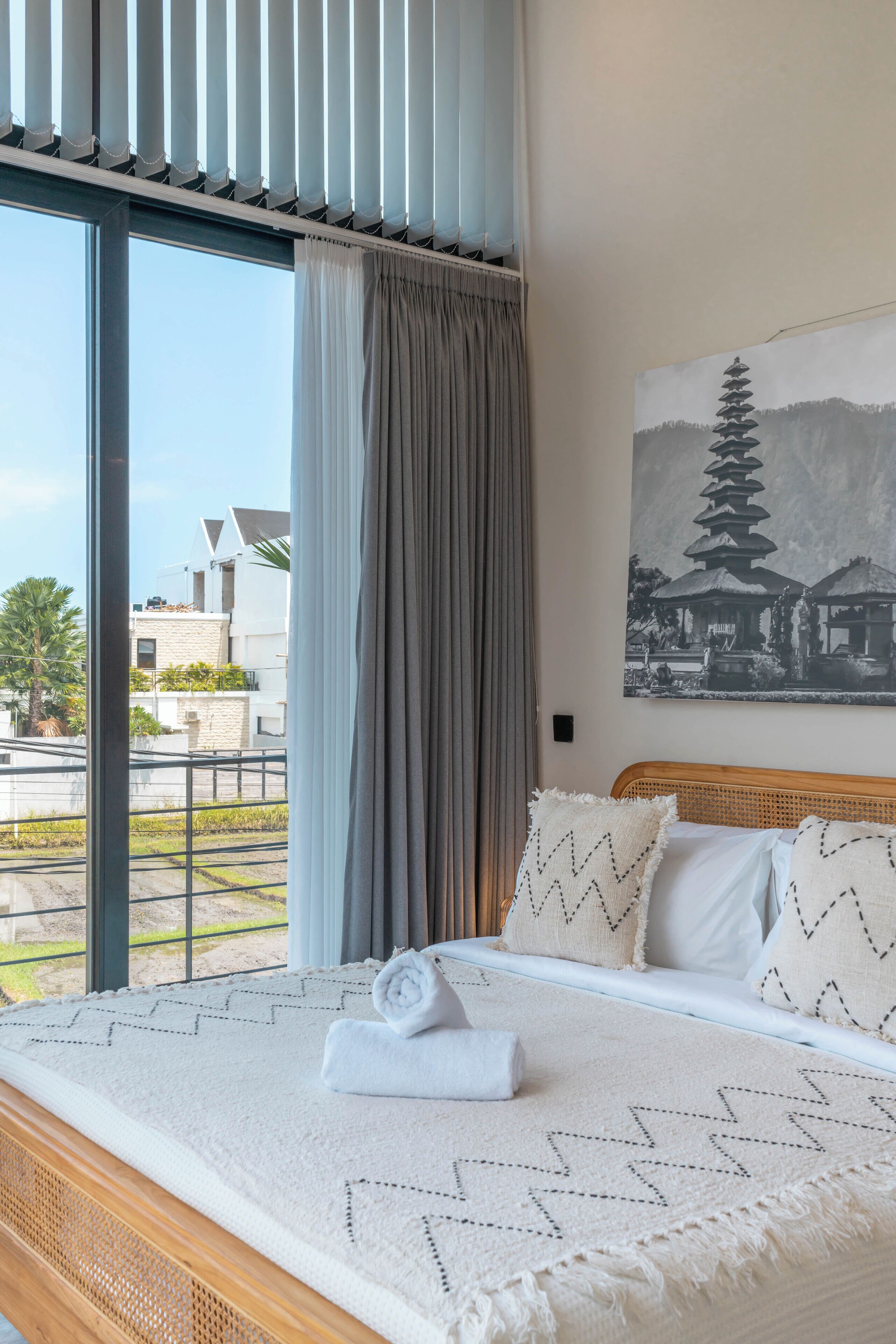 A neatly made bed with a patterned blanket and pillows, a large window with curtains, and a wall-mounted artwork of a pagoda.