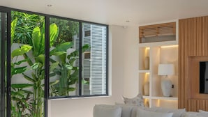 A modern living room with a white sofa, a large window with blinds, a wooden shelf with decorative items, and a view of greenery outside. - Villa Maverick (Canggu)