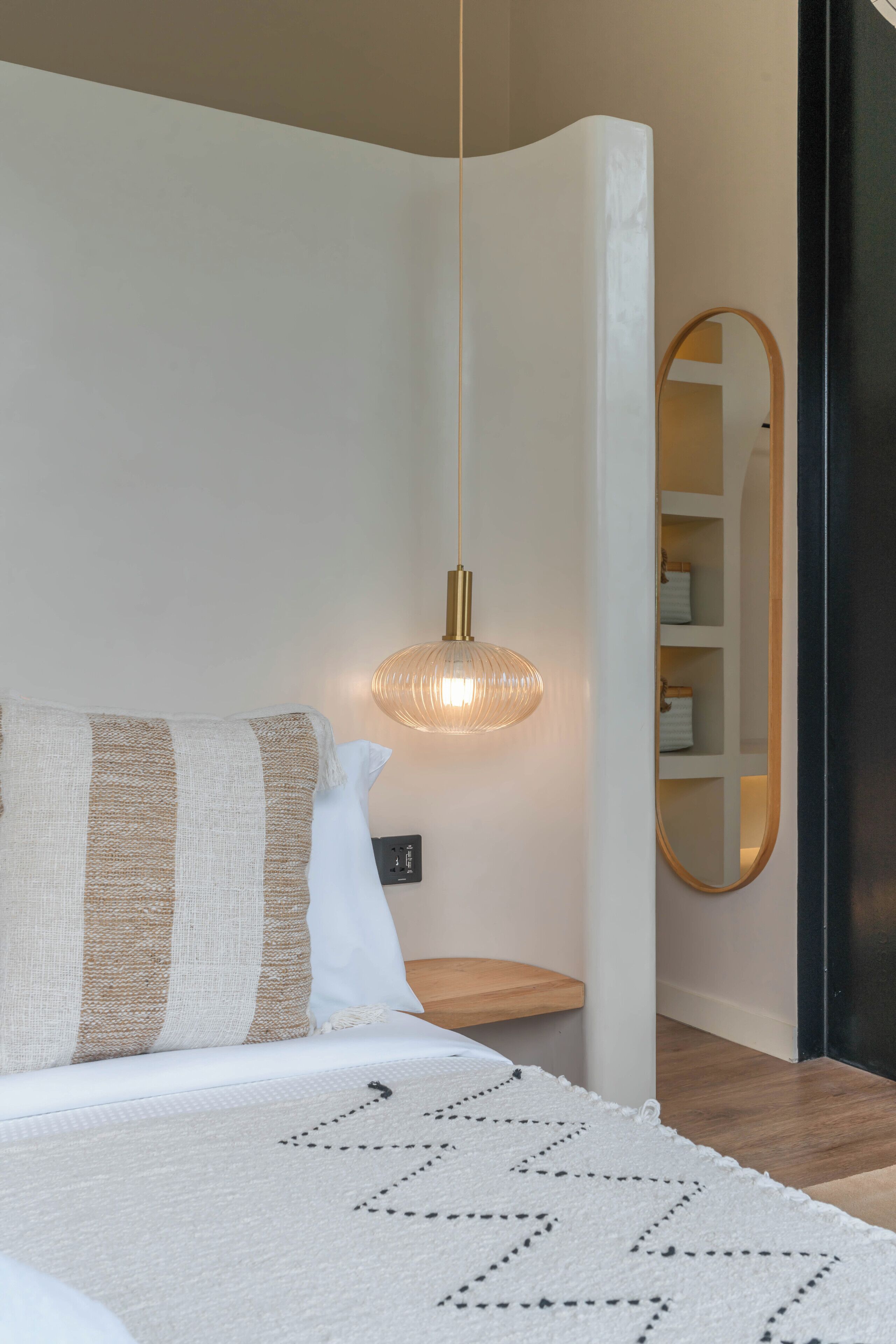 A bedroom with a bed, a bedside lamp, a mirror, and a wooden shelf.