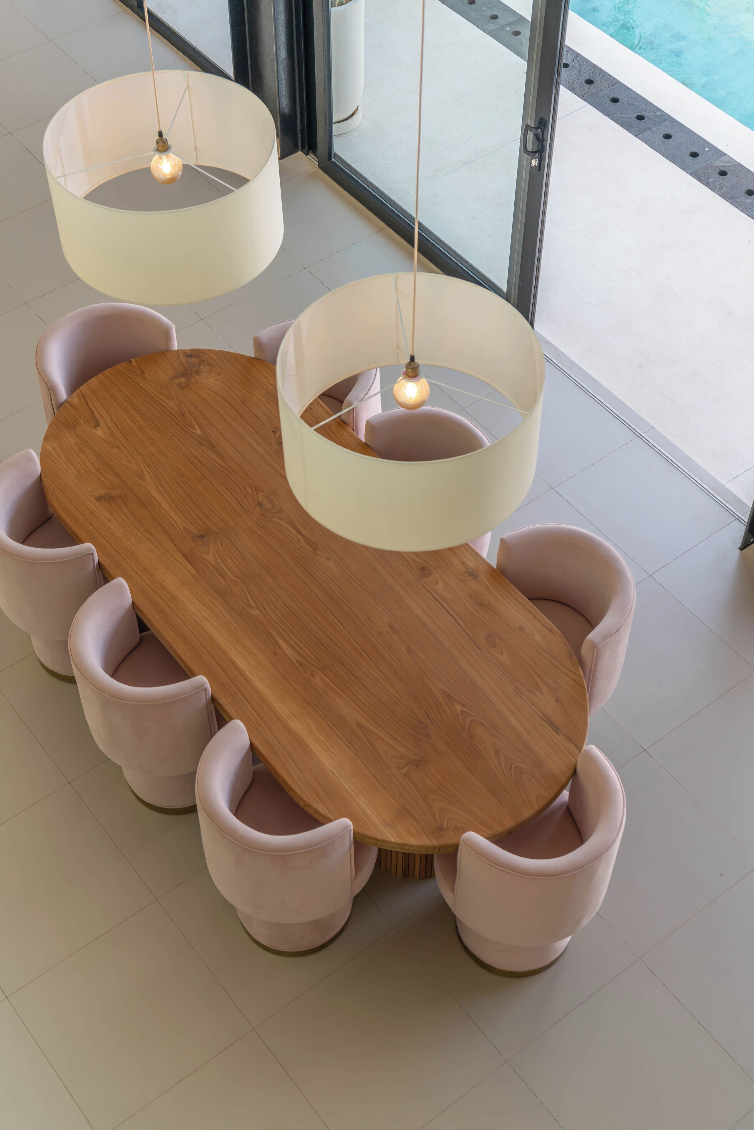 Una mesa redonda de madera con seis sillas acolchadas, dos lámparas colgantes y vistas a una piscina a través de puertas de cristal.
