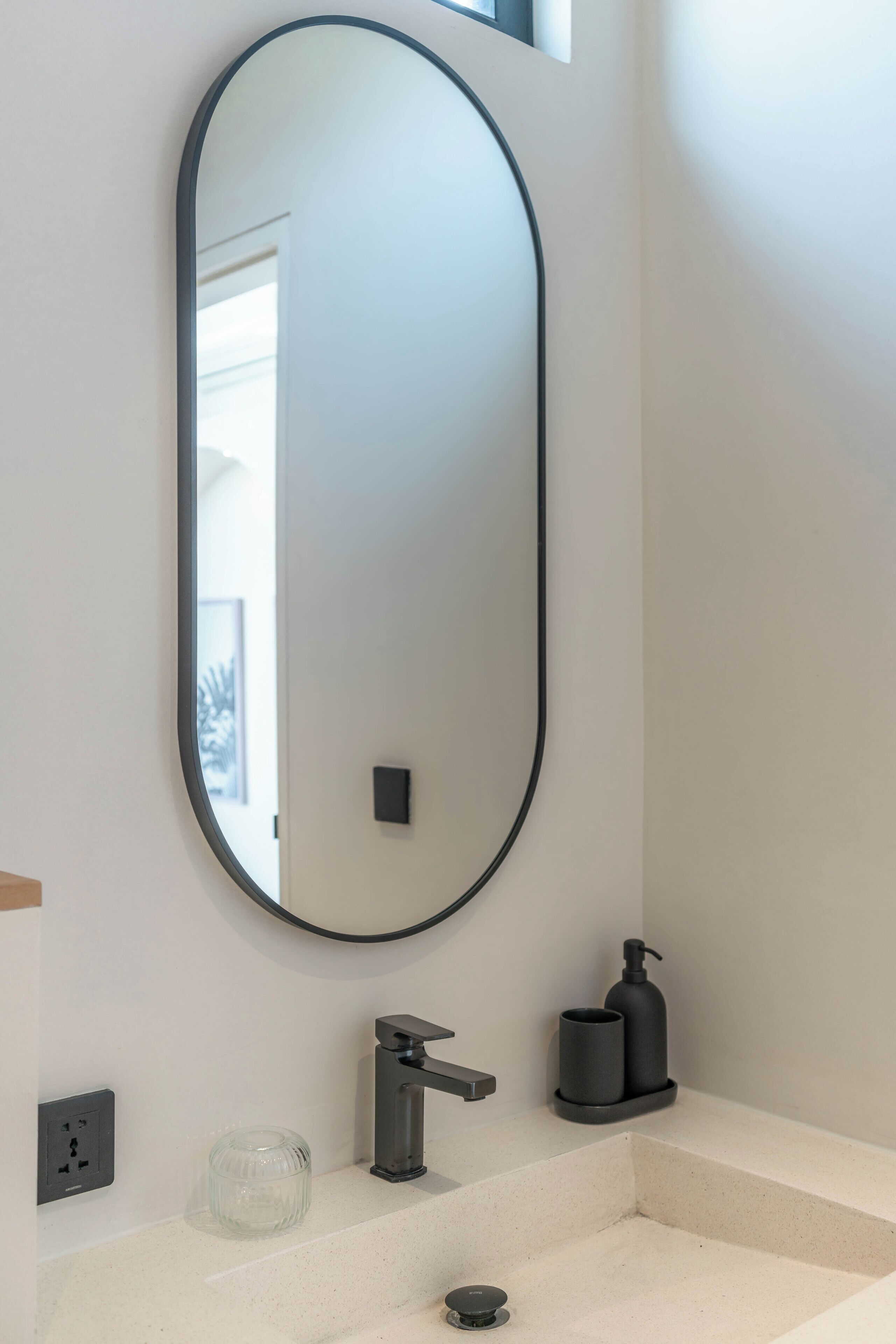 A bathroom with a large oval mirror, a modern faucet, a glass jar, and a soap dispenser.