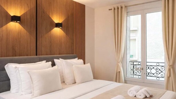 A hotel room with a bed, pillows, a headboard, wall-mounted lights, a window with curtains, and a balcony.
