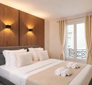 Ein Hotelzimmer mit Bett, Kissen, Kopfteil, Wandleuchten, einem Fenster mit Vorhängen und einem Balkon.