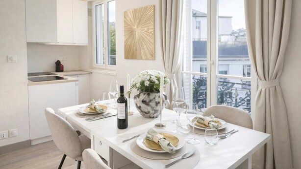 A modern dining area with a white table set for two, a vase with flowers, and a bottle of wine.