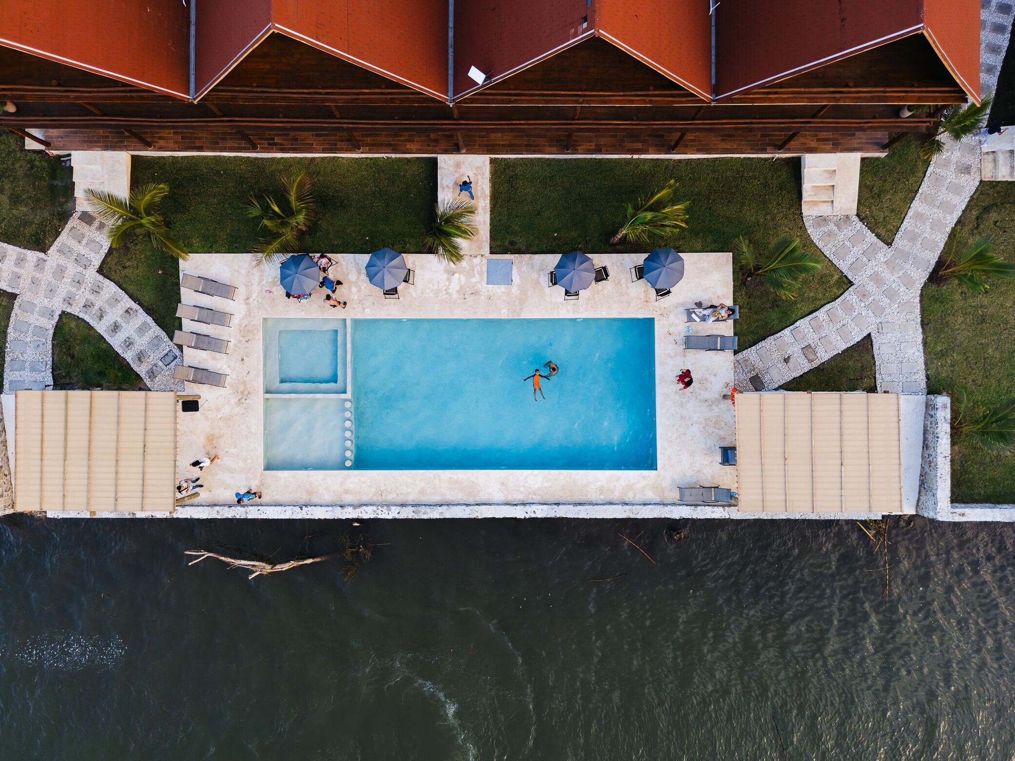 An aerial view of a rectangular swimming pool with a small hot tub, surrounded by a deck with umbrellas and lounge chairs.
