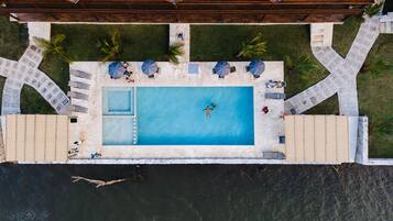 An aerial view of a rectangular swimming pool with a small hot tub, surrounded by a deck with umbrellas and lounge chairs.