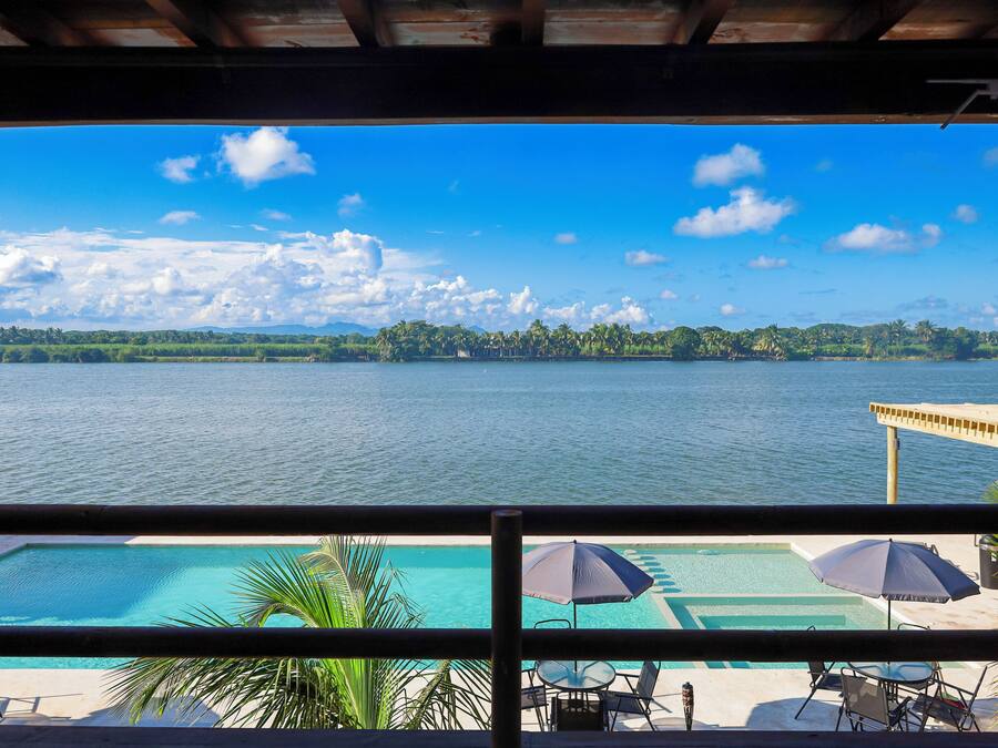 A view of a swimming pool with umbrellas and chairs, overlooking a large body of water and a forested shoreline.
