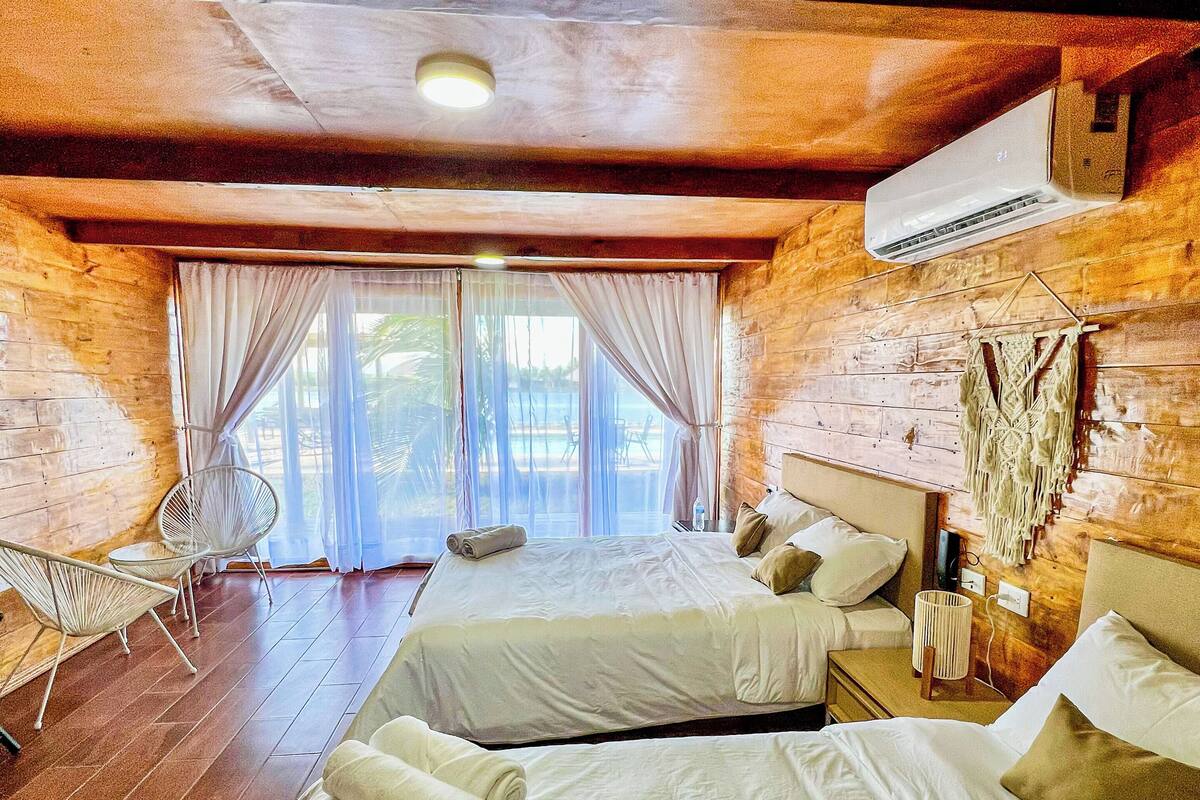 Ein Zimmer mit zwei Betten, Holzboden, einem großen Fenster mit Vorhängen und einer hängenden Wandweberei aus Makramee.
