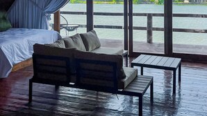 An attic room with a bed, a sofa, and a table, overlooking a body of water and greenery.