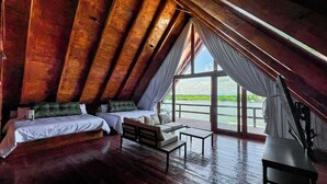 A cozy attic bedroom with two beds, a sofa, and a table, featuring a large window with a view of a body of water.