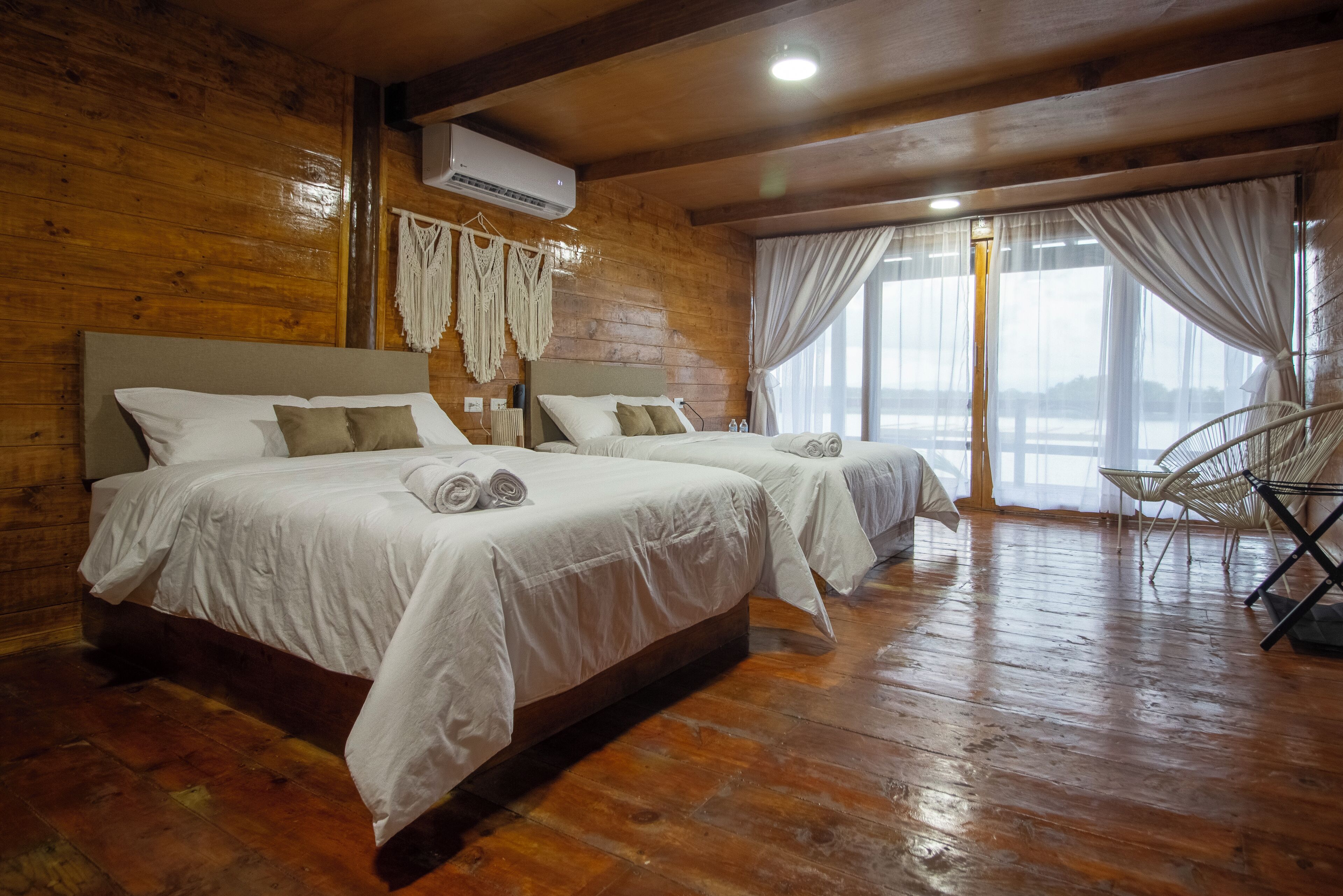 Una habitación tipo cabaña de madera con dos camas, un ventanal con cortinas y una silla.