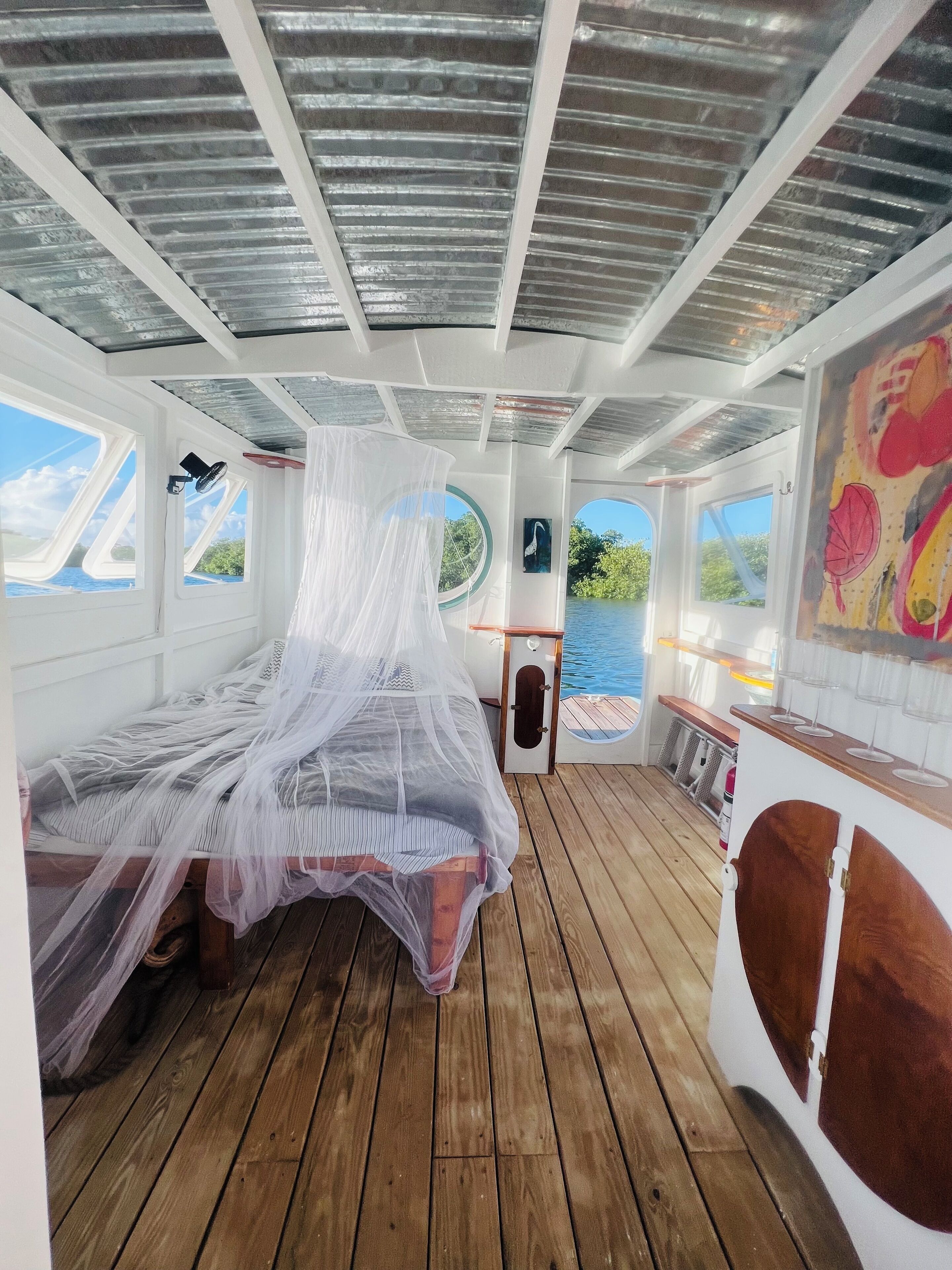 Una cabina con un letto coperto da una zanzariera, pavimento in legno e ampie finestre con vista sull'acqua.