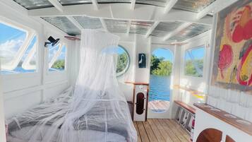 Una cabina con un letto coperto da una zanzariera, pavimento in legno e ampie finestre con vista sull'acqua.