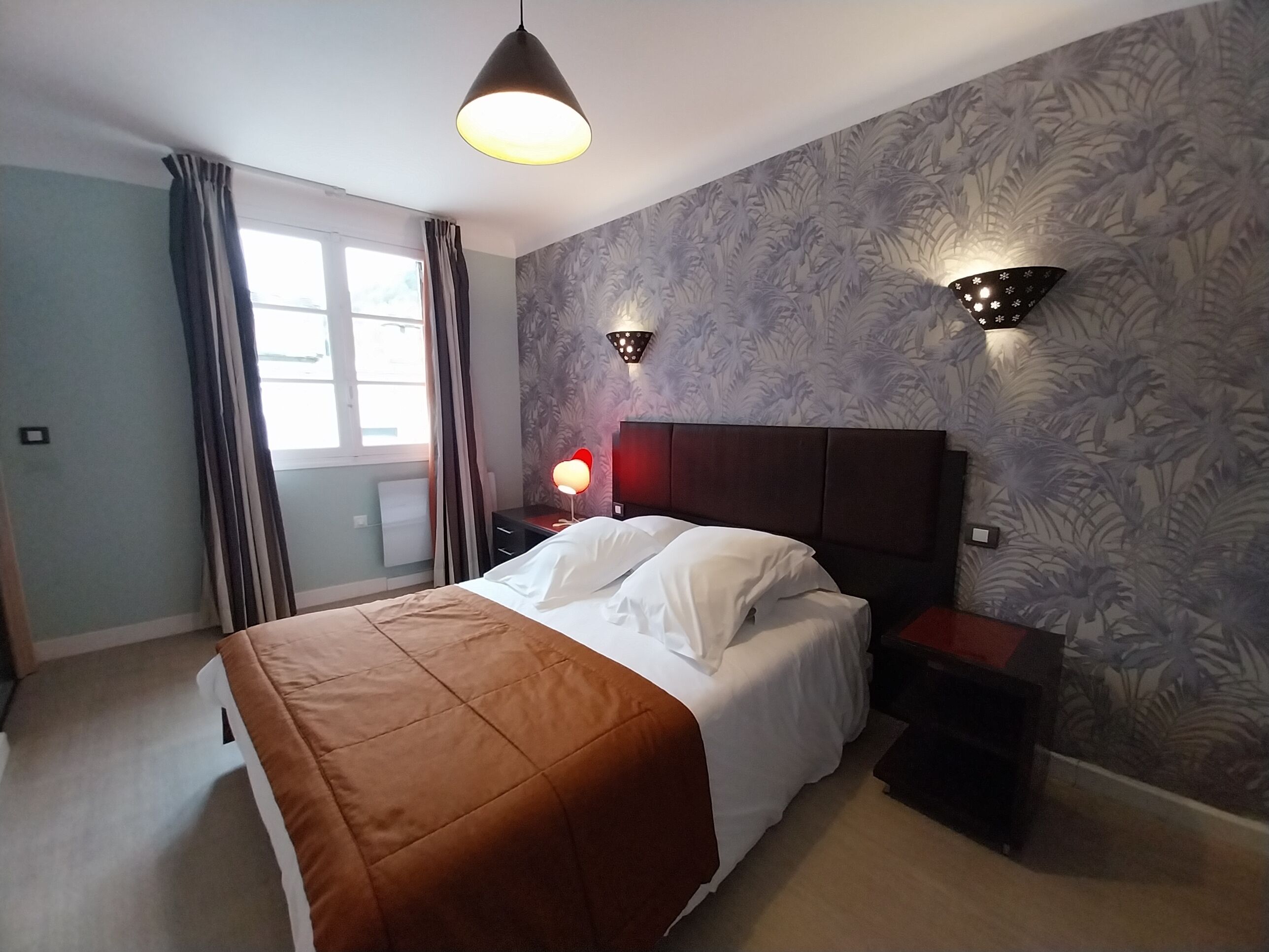 Un dormitorio con cama, mesita de noche, lámpara de pared, ventana con cortinas y un papel tapiz estampado.