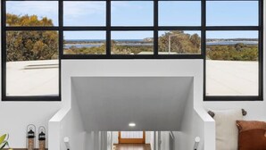 A spacious interior with a high ceiling, large windows offering views of a natural landscape, and a central staircase leading to an upper level. - Luxurious Beach House with Breath-taking Views (Ocean Grove)
