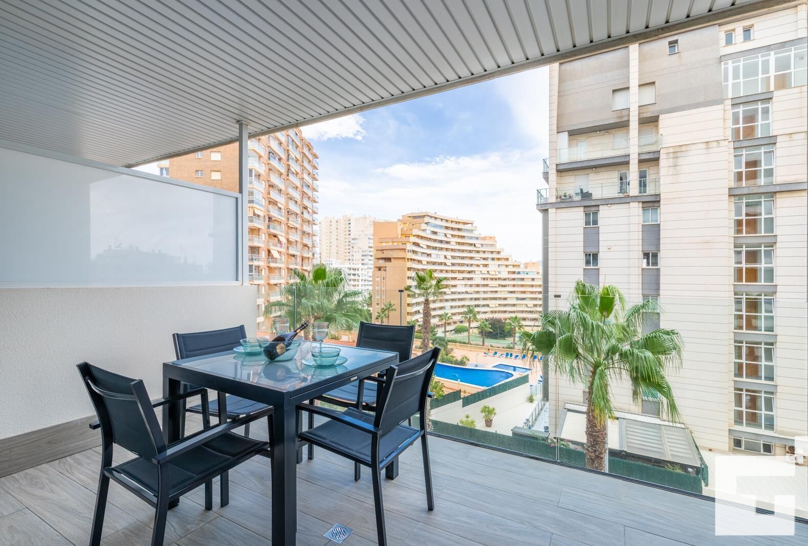 Un balcón con mesa y sillas, con vista a una piscina y edificios altos.