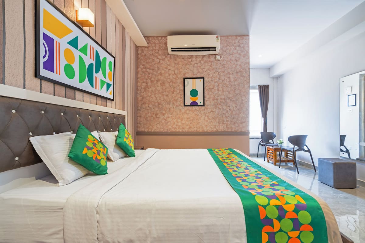 A hotel room with a bed, a headboard, a wall-mounted air conditioner, a framed picture, and a seating area with chairs and a table.