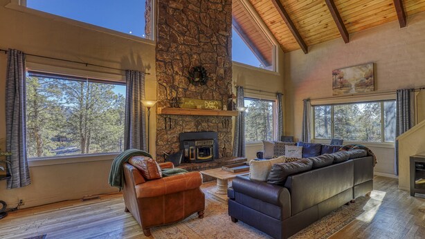 A spacious living room with a stone fireplace, leather sofas, and large windows offering a view of the trees outside.