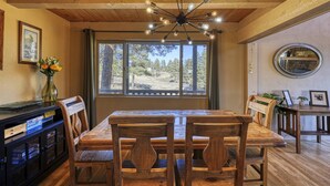 A dining area with a wooden table and chairs, a chandelier, a window with a view of trees, and a mirror on the wall.