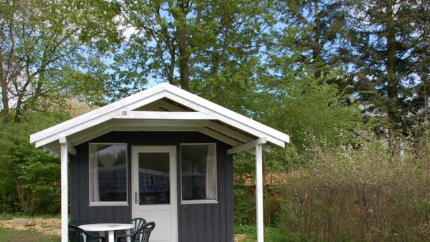 A small, dark-colored wooden structure with a white roof, a door, and two windows. It has a table and chairs outside. - 2 Person Holiday Park Home in Jelling (Jelling)