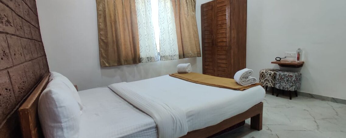Uma cama de solteiro com estrutura de madeira e roupa de cama branca, uma pequena mesa de cabeceira com uma toalha e uma janela com cortinas.