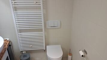 A small bathroom with a toilet, a radiator, a trash bin, and a toilet paper holder.