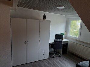 Une pièce avec une armoire blanche, un bureau et une chaise, et une fenêtre qui laisse entrer la lumière naturelle.