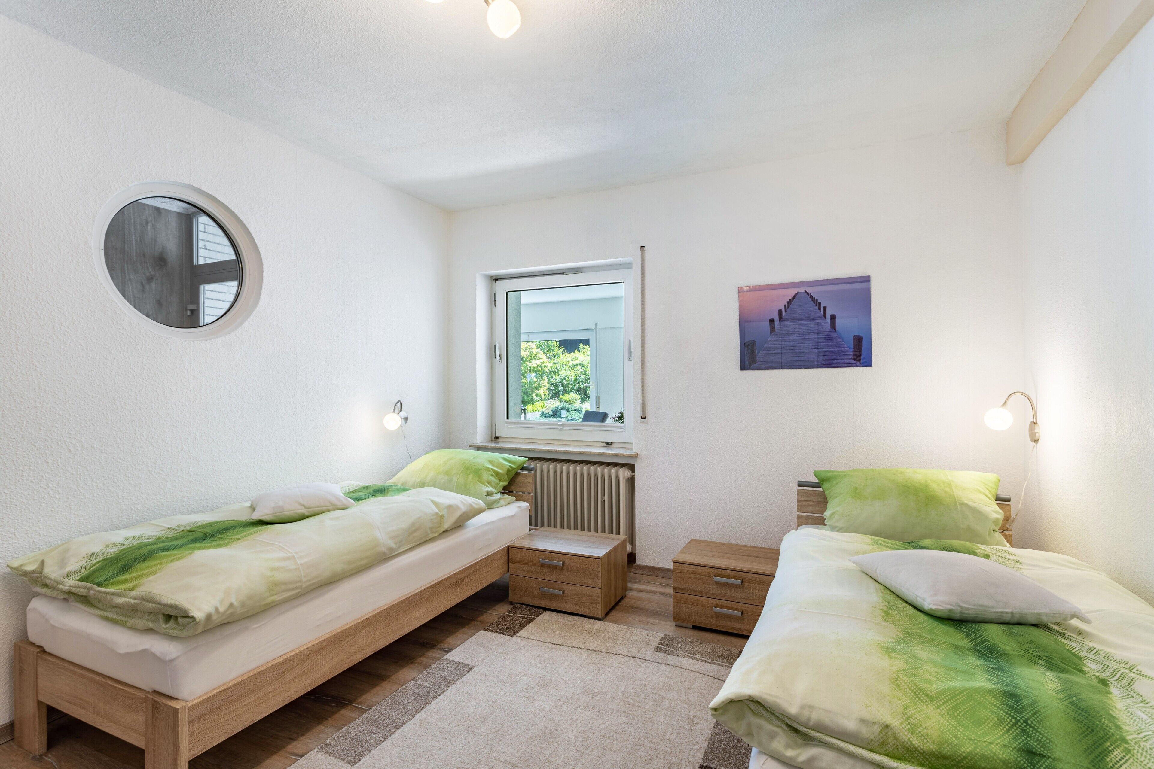 A bedroom with two single beds, a round mirror, a window with a view, and a radiator.