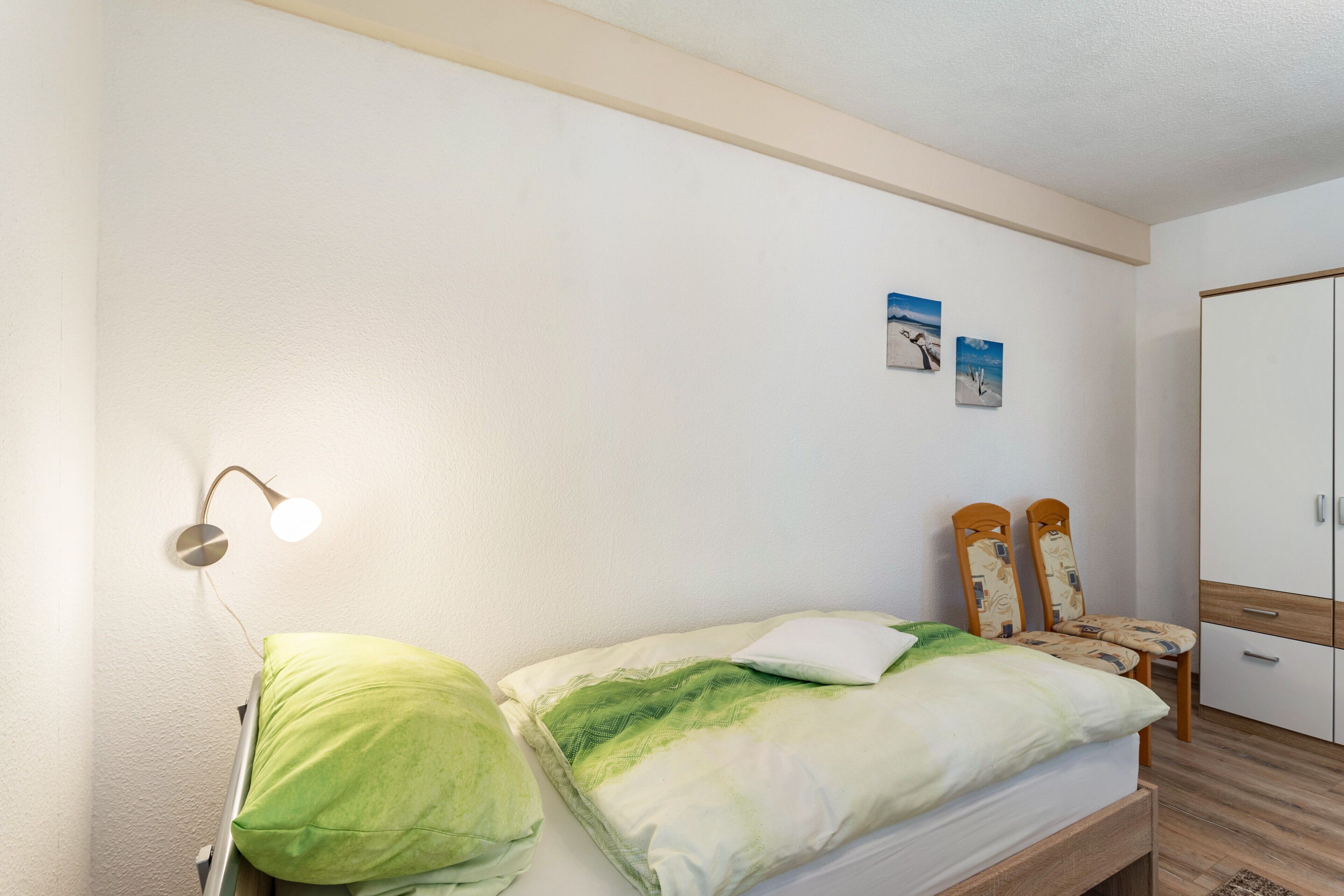 Una cama individual con ropa de cama verde y blanca, una lámpara de noche, una mesita con una silla y un armario.