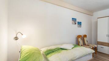 Una cama individual con ropa de cama verde y blanca, una lámpara de noche, una mesita con una silla y un armario.