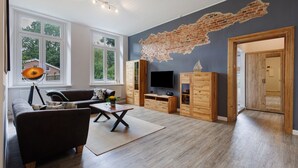 A living room with a large window, a flat-screen TV, wooden cabinets, and a brick wall mural. - Apartment 'Reit- Und Ferienhof Alte Dorfschule' with  Private Terrace and Wi-Fi (Holstenniendorf)